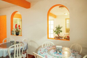 A dining area with a round table and chairs, two arched windows, and a view of a room with a plant and window blinds. - OYO Hotel Meson Del Rio (Tepotzotlán)