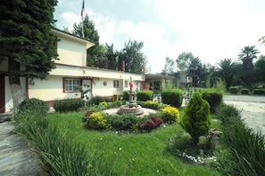 A building with a garden featuring a fountain, neatly trimmed hedges, and a flag. - Capital O Hotel Meson Del Rio, Tepotzotlan (Tepotzotlán)