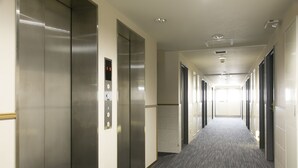 A long, narrow hallway with a carpeted floor, flanked by doors on both sides, leading to an elevator on the left. - Ace Inn Matsumoto (Matsumoto)