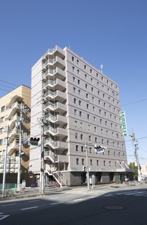 Un hotel de varios pisos con diseño moderno, que cuenta con balcones en cada planta, una entrada visible y un cielo despejado al fondo.