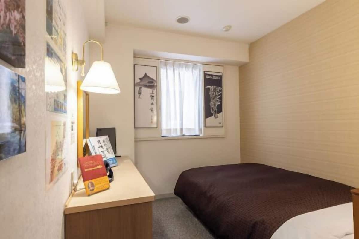 Ein kleines, ordentlich eingerichtetes Hotelzimmer mit einem Einzelbett, einem Schreibtisch mit Lampe und einem Fenster mit Vorhängen.
