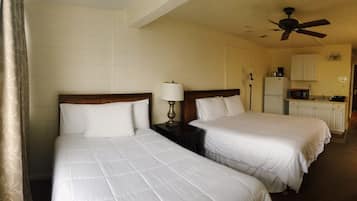 A hotel room with two beds, a ceiling fan, a small table with a lamp, a refrigerator, and a microwave.