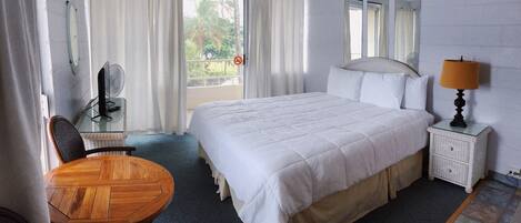 A hotel room with a large bed, a wooden dining table, a chair, a nightstand with a lamp, a fan, and a view of palm trees through the window.