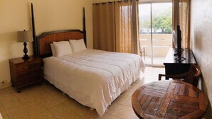 A hotel room with a bed, a wooden nightstand, a desk with a chair, a round wooden table, and a ceiling fan. - Hilo Reeds Bay Hotel (Hilo)