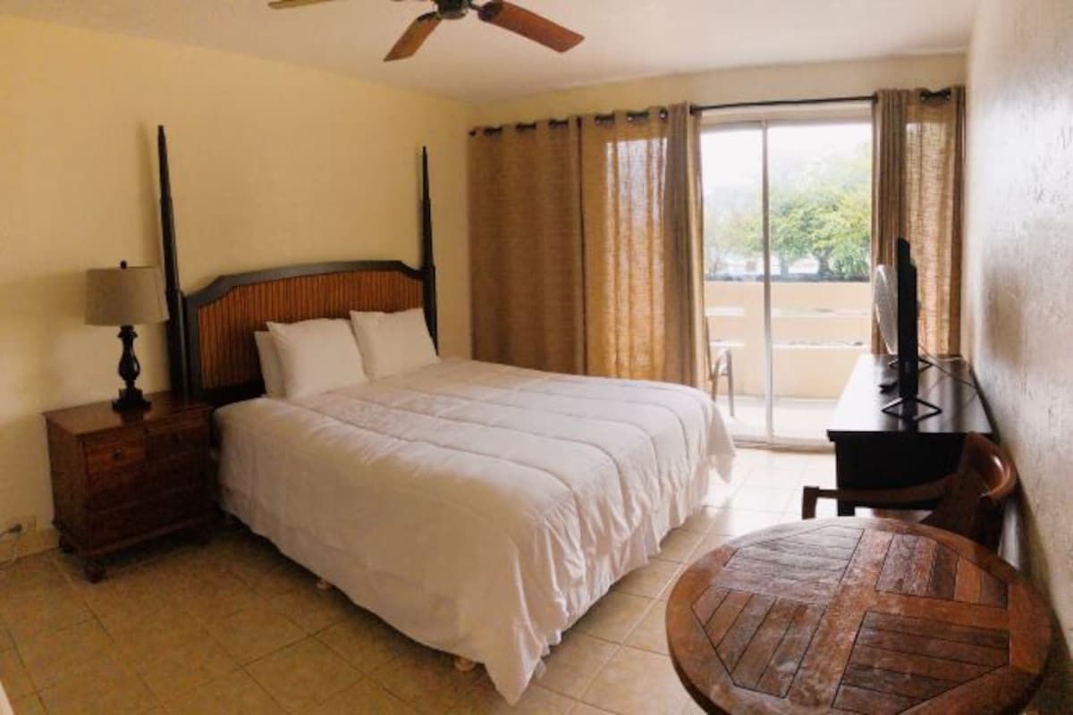 A hotel room with a bed, a wooden nightstand, a desk with a chair, a round wooden table, a ceiling fan, and a view of the outdoors through a sliding door.