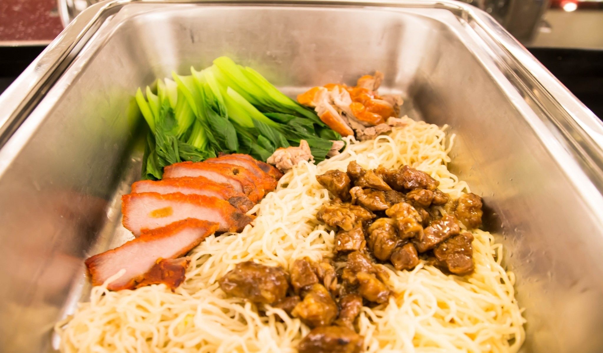 A stainless steel food container with sliced meat, noodles, and green vegetables.