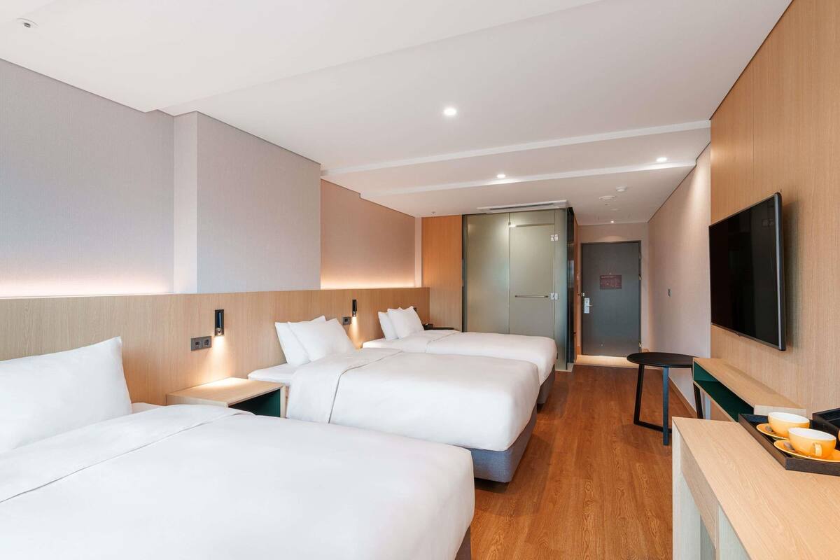 A hotel room with three beds, a flat-screen TV, a wooden desk, and a door leading to another room.