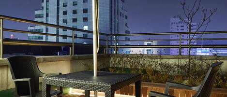 An outdoor seating area with a table and chairs under a large umbrella, overlooking a cityscape at night.