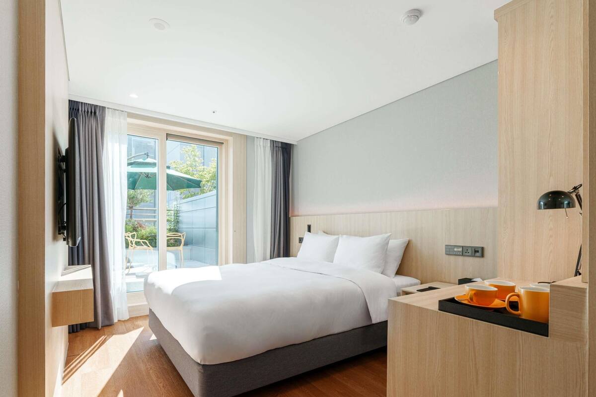 A modern hotel room with a large bed, a desk with a lamp, and a view of the outdoors.