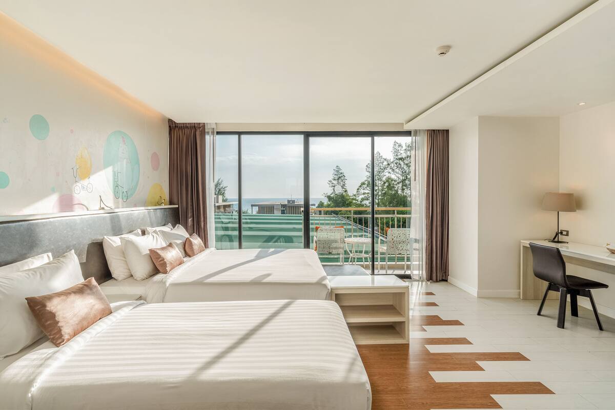 A hotel room with two beds, a desk, and a view of the sea.