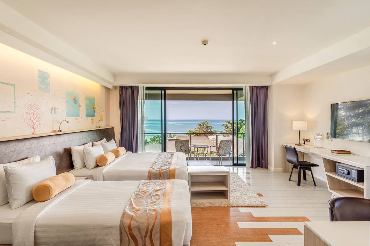 A hotel room with two beds, a desk, a chair, and a balcony with a view of the sea.