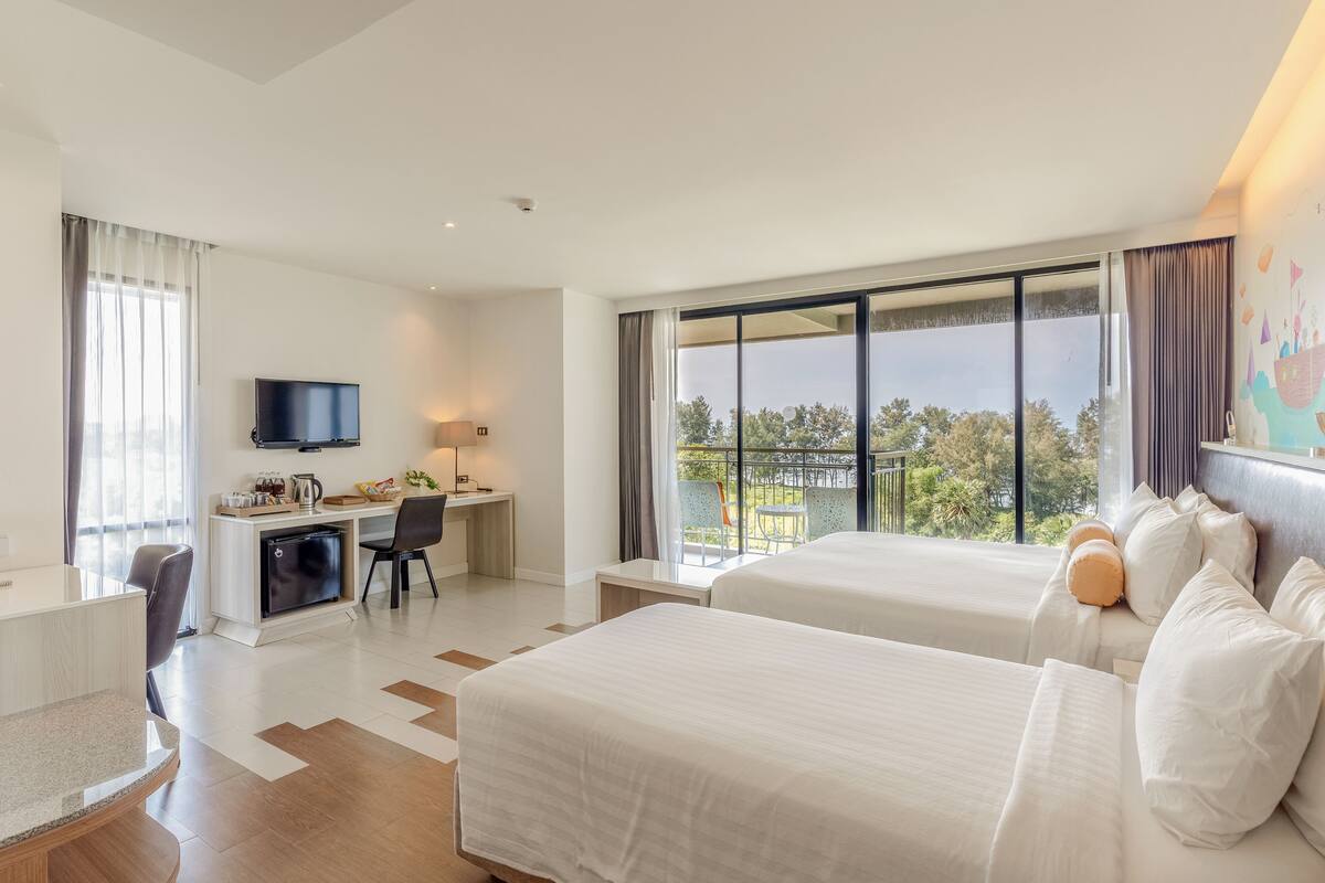 A hotel room with two beds, a desk, a TV, and a balcony with a view of trees.