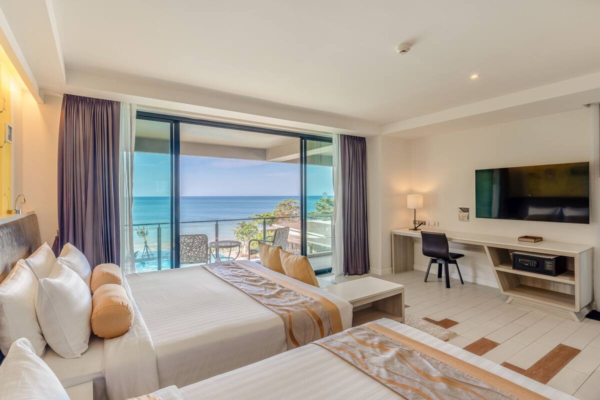 A hotel room with two beds, a desk, a TV, and a balcony with a view of the ocean.