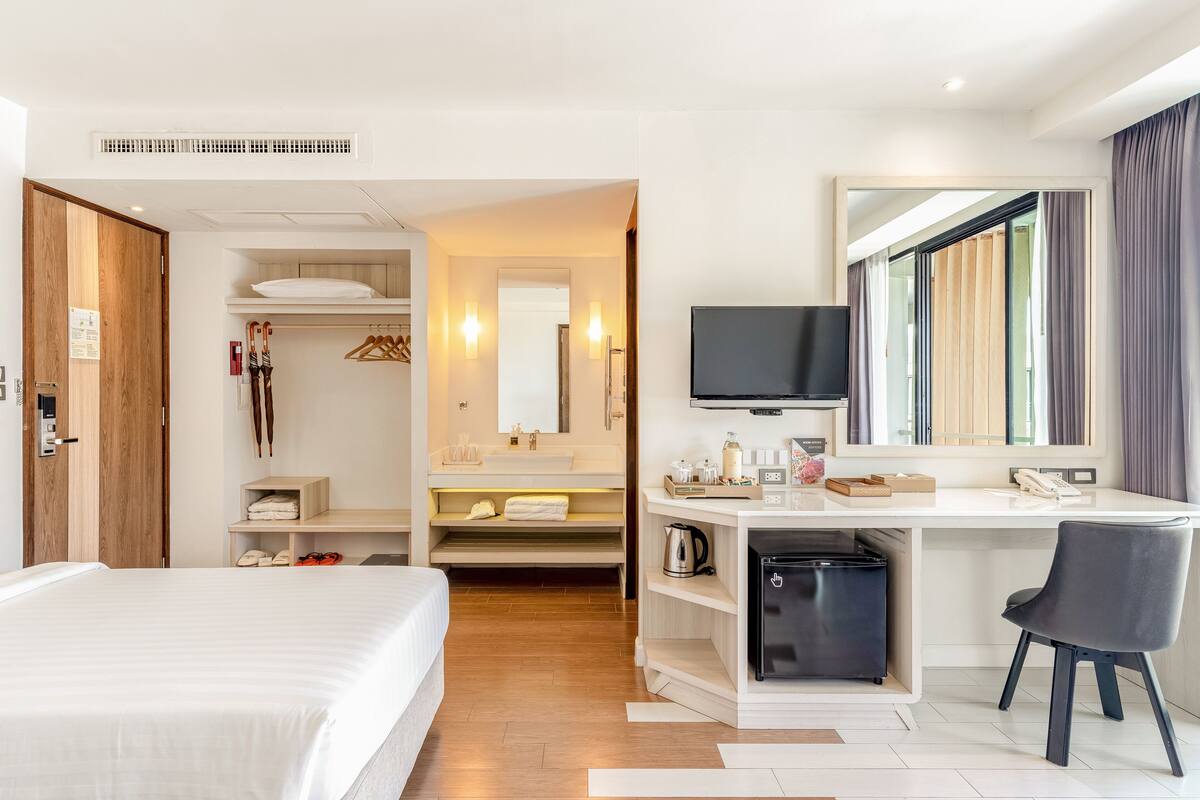 A hotel room with a bed, a desk with a chair, a television, and a bathroom with a mirror and towel rack.