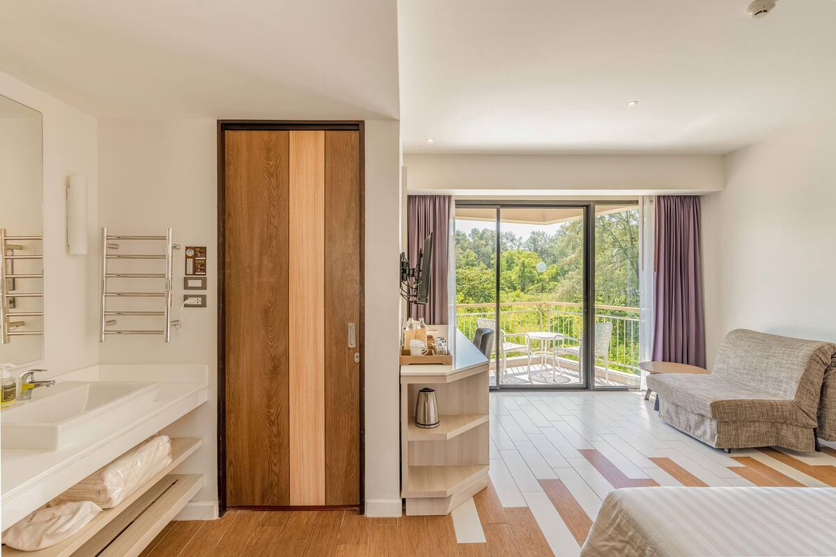 A modern bedroom with a wooden door, a white sink, a bed, a sofa, and a balcony with a view of greenery.