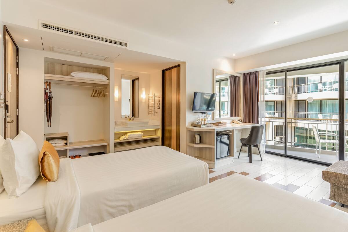 A hotel room with two beds, a desk, a TV, and a balcony with a view of the city.