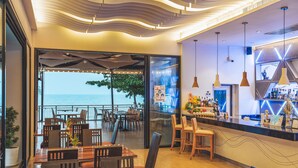A modern restaurant with a view of the sea, featuring wooden tables and chairs, a bar area, and a ceiling with a wave-like design. - Sand Dunes Chaolao Beach Resort (Tha Mai)