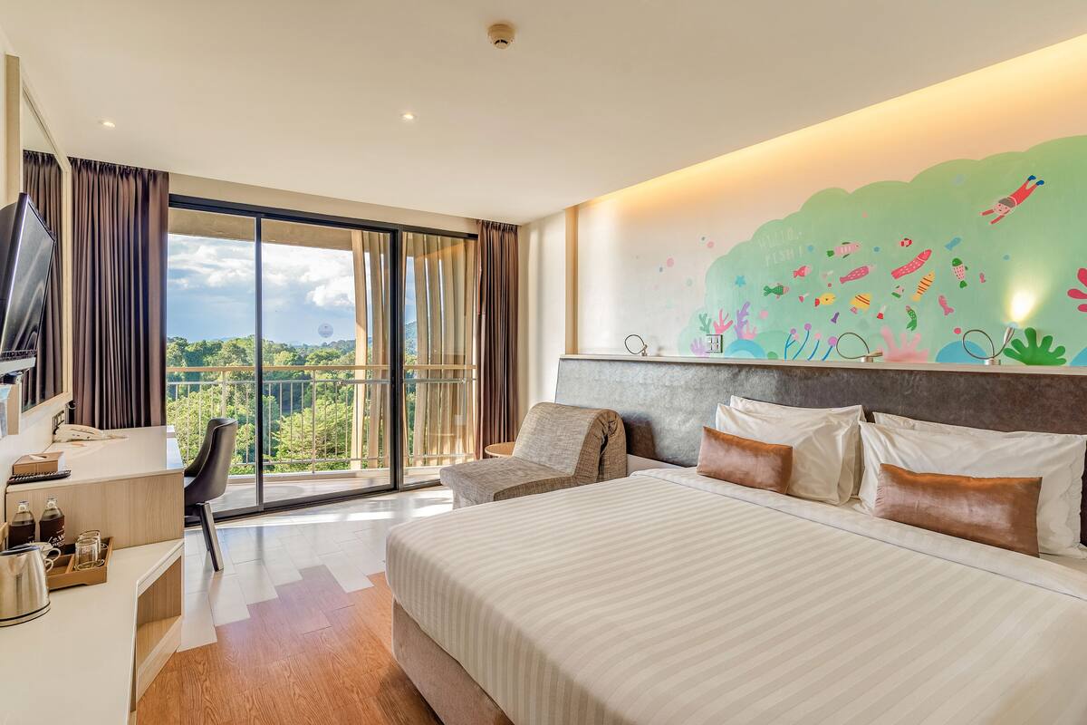 A hotel room with a large bed, a desk with a TV, and a view of greenery through the window.