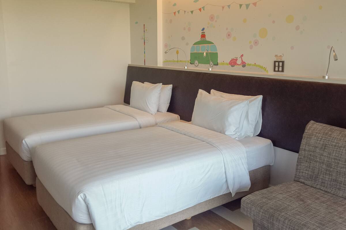 A hotel room with two beds, a dark headboard, a small armchair, and a wall with colorful decorations.