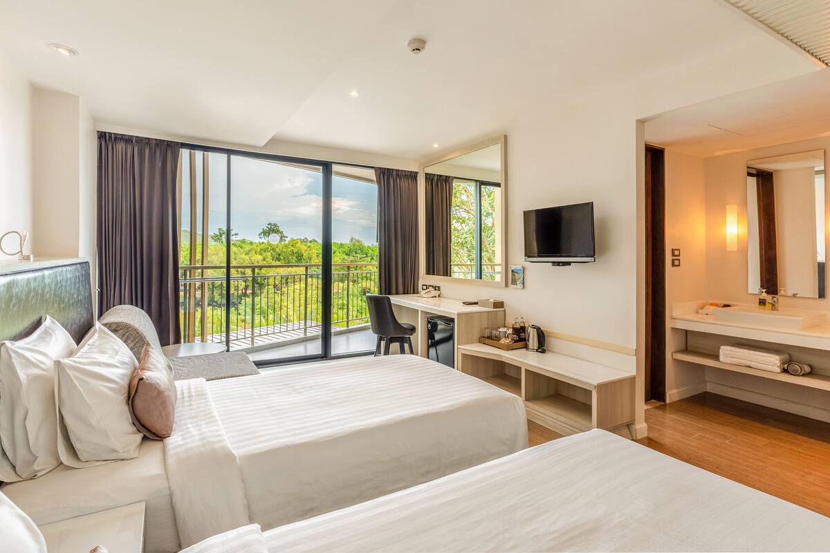 A modern hotel room with two beds, a desk, a computer, a TV, and a balcony with a view of greenery.