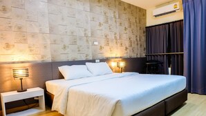 A neatly made bed with white linens, a bedside table with a lamp, and a wall with a patterned design. - Prajaktra City Hostel (Udon Thani)