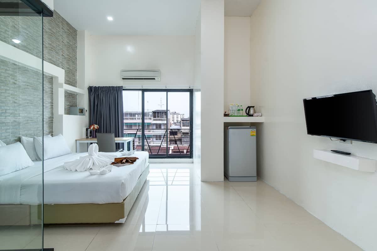 A modern hotel room with a bed, a desk, a chair, a TV, and a view of the city through large windows.