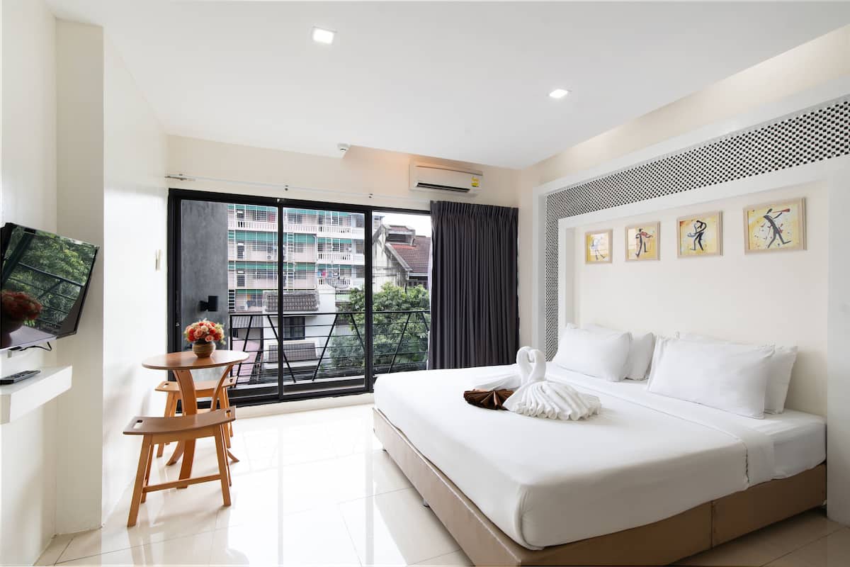 A modern hotel room with a large bed, a desk with a chair, a TV, and a balcony with a view of buildings.