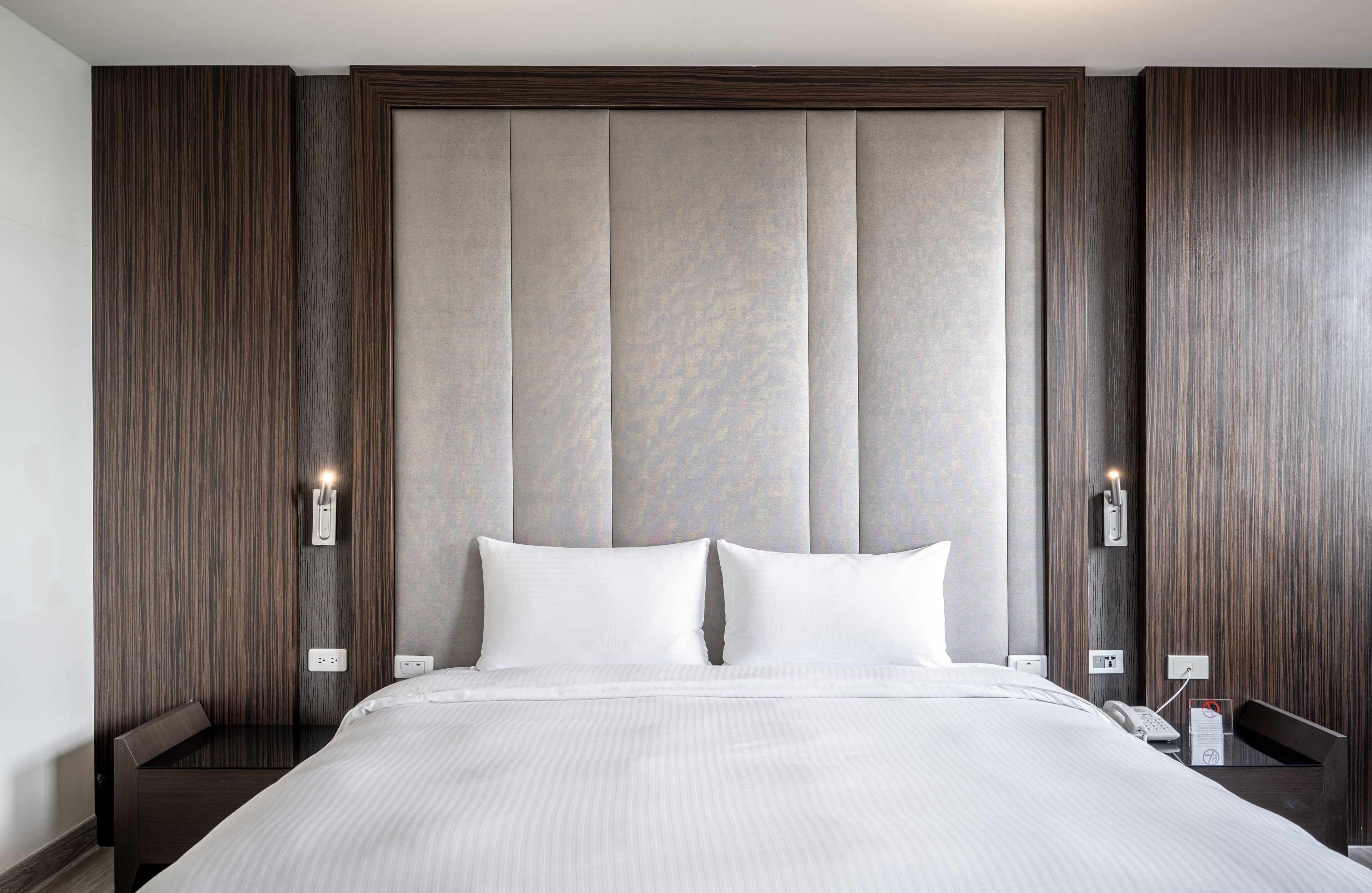Habitación de hotel con una cama grande, paneles de madera en las paredes y cabecera en tonos neutros. Se observan mesitas de noche con lámparas y un teléfono a un lado.