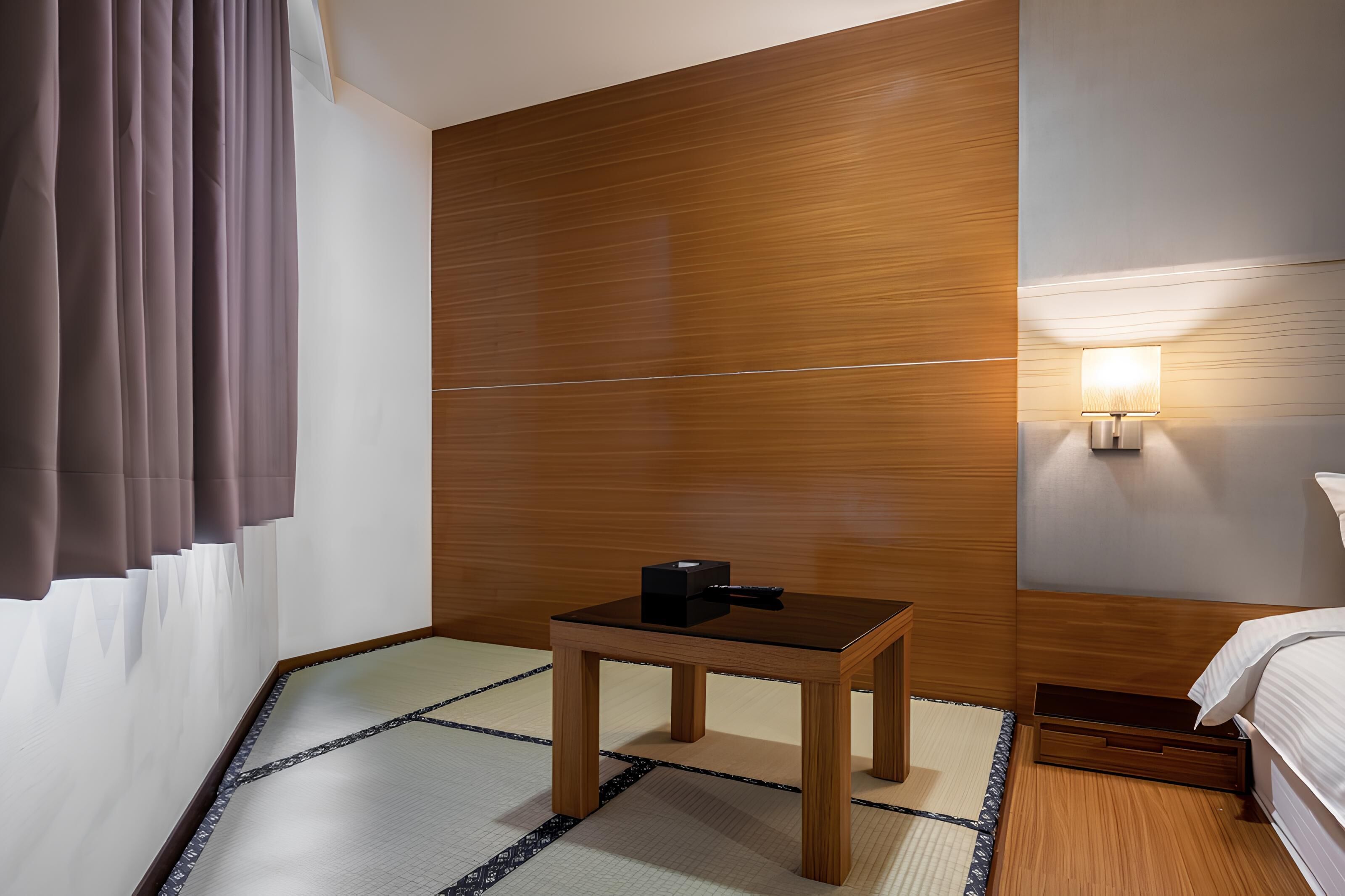A hotel room with a wooden panel wall, a small wooden table with a black object, a bedside lamp, and a bed with white bedding.