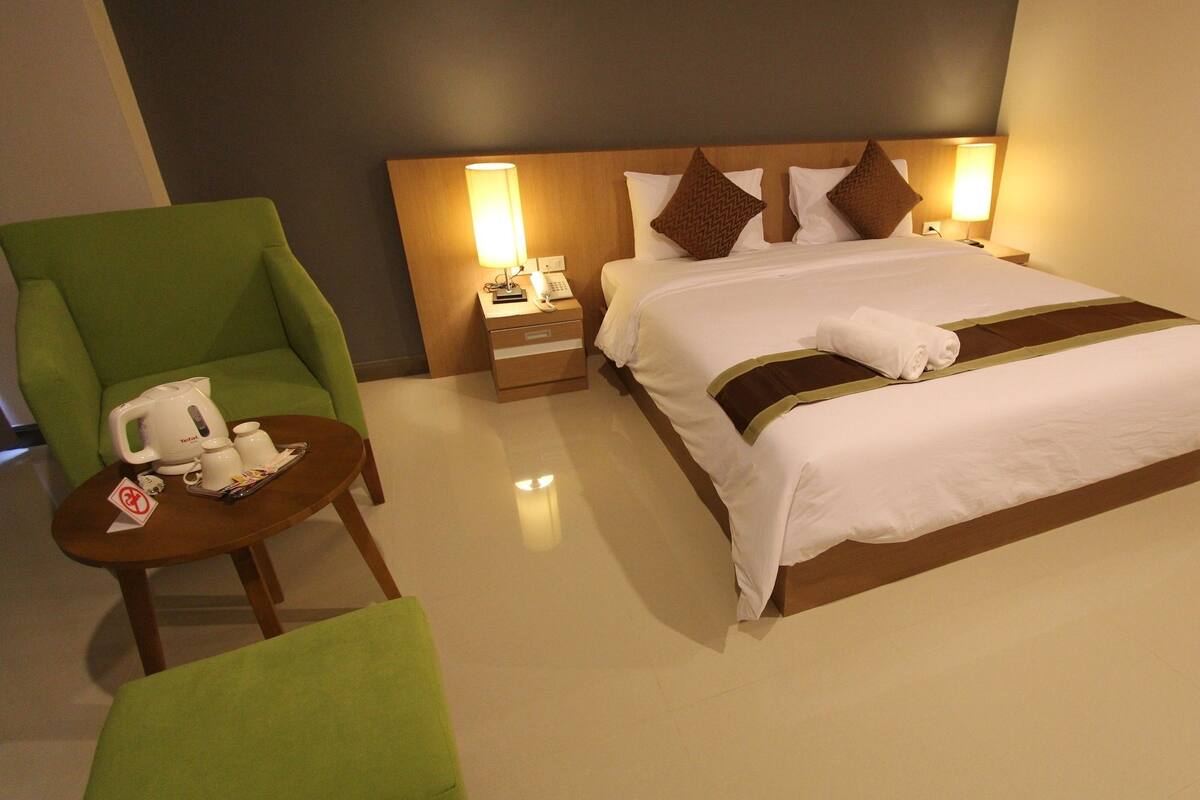 A hotel room with a large bed, two bedside lamps, a green armchair, a small table with a kettle and cups, and a telephone.