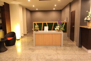 A reception desk with a wooden counter, a green decorative panel, and a vase with flowers. There is a wicker chair with a cushion and a lamp to the left. - S Tara Grand Hotel (Surat Thani)