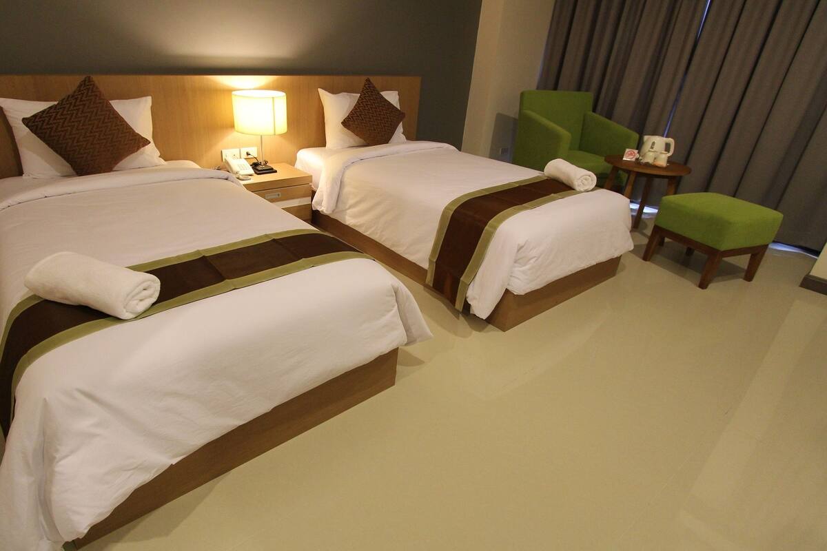 A hotel room with two beds, a nightstand with a lamp, a telephone, a green chair, and a small table with a coffee cup.