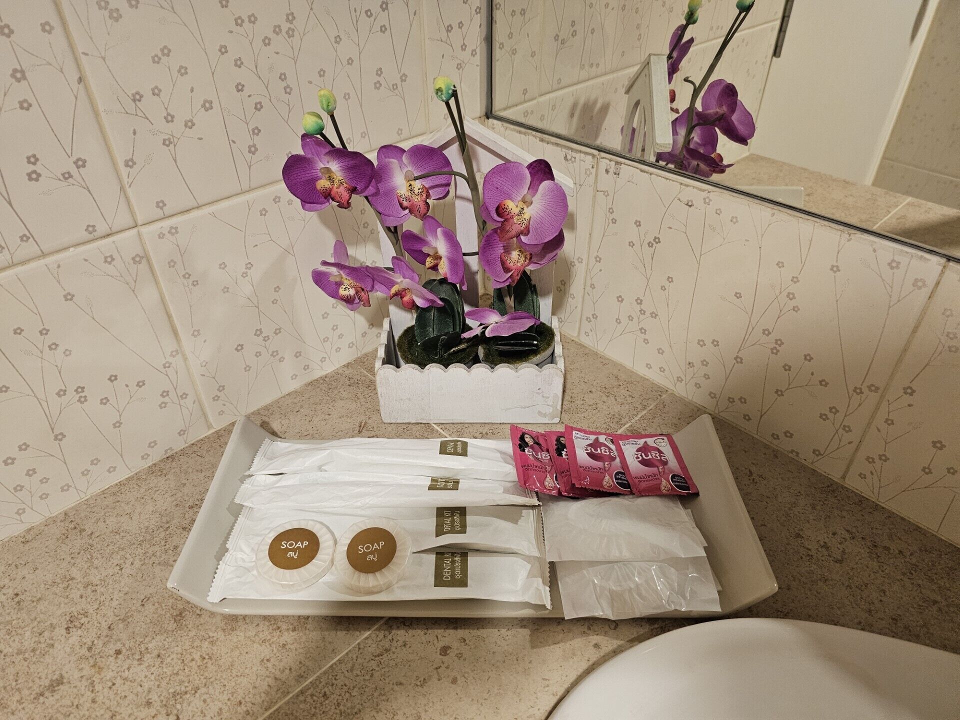 A bathroom with a floral tile backsplash, a white sink, a mirror, and a small orchid plant in a white pot. There are also two soap bars and a pack of pink soap bars on the counter.