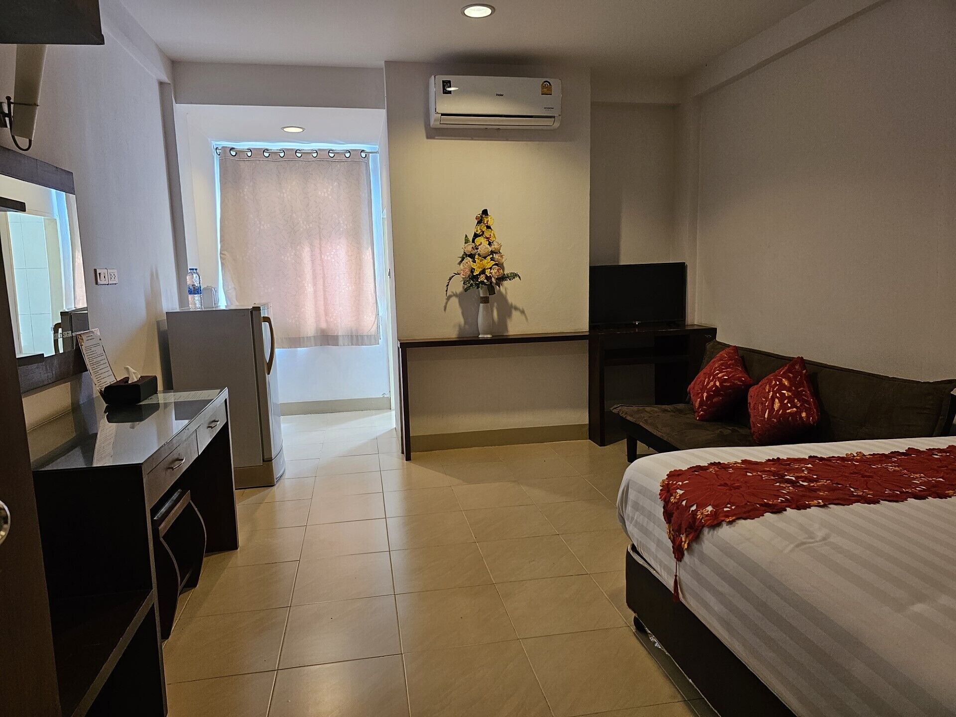 Ein Hotelzimmer mit Bett, Schreibtisch, Fernseher, einem kleinen Tisch mit Blumenvase und einer Klimaanlage.