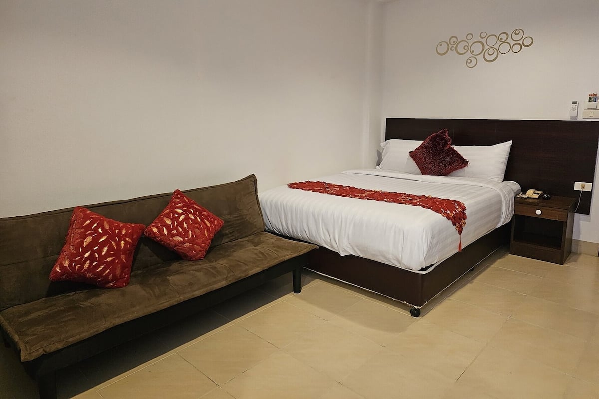 A hotel room with a bed, a brown sofa with red pillows, a bedside table with a phone, and a wall decoration.