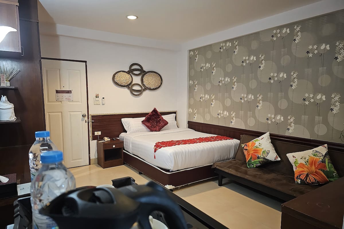 A hotel room with a bed, bedside table, and a sofa. There are water bottles and a kettle on the table.