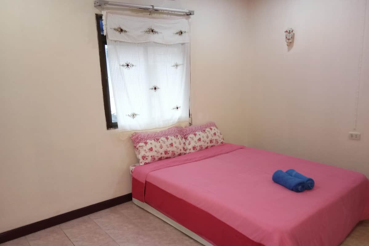 A bedroom with a bed, a window with a curtain, a wall clock, and a power outlet.
