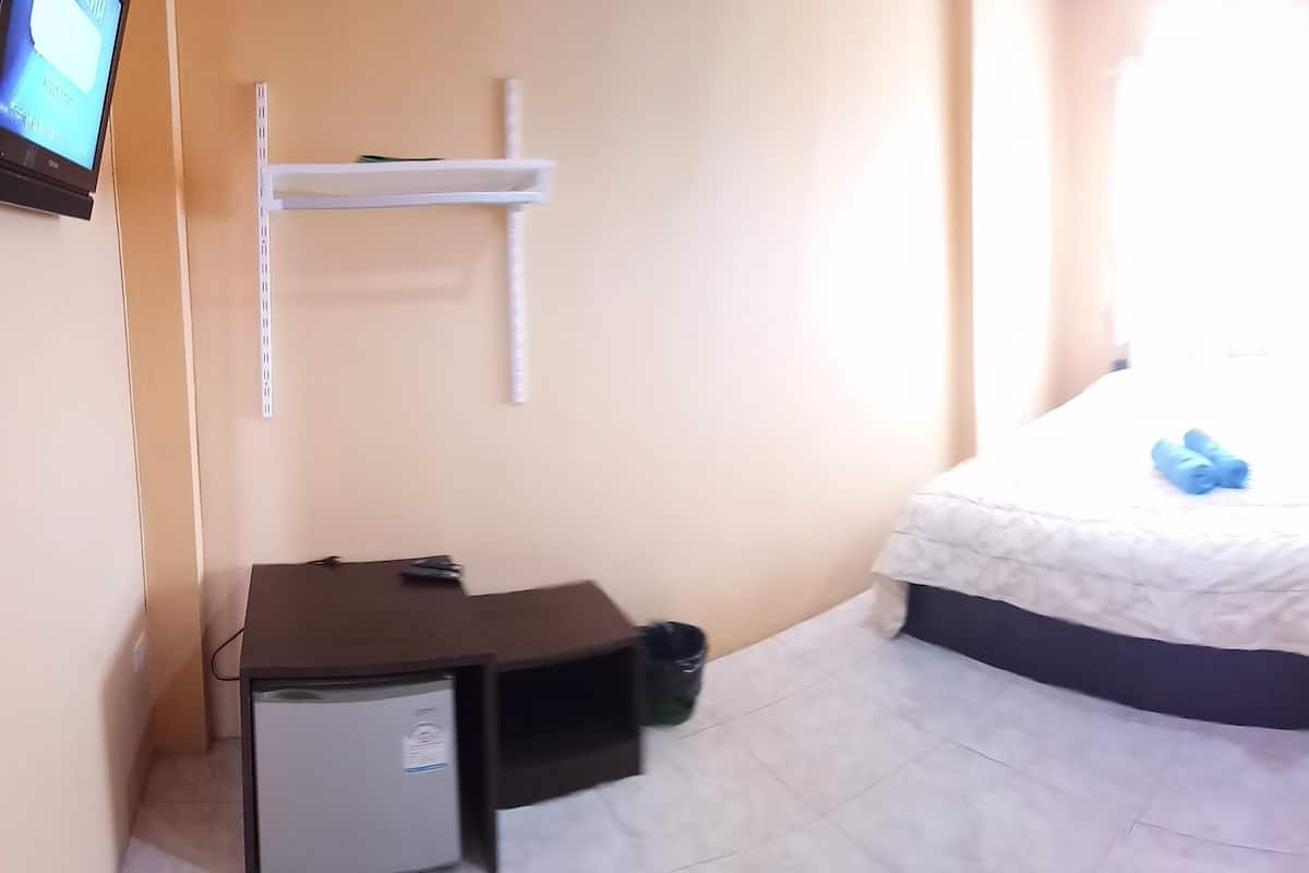A small, well-lit room with a bed, a desk, a chair, and a wall-mounted shelf.