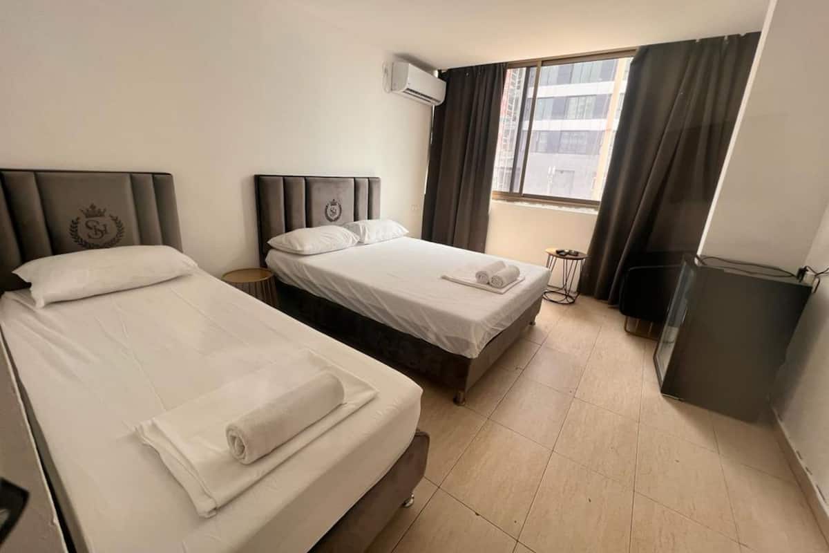 A hotel room with two beds, a small table, an air conditioning unit, and a window with curtains.