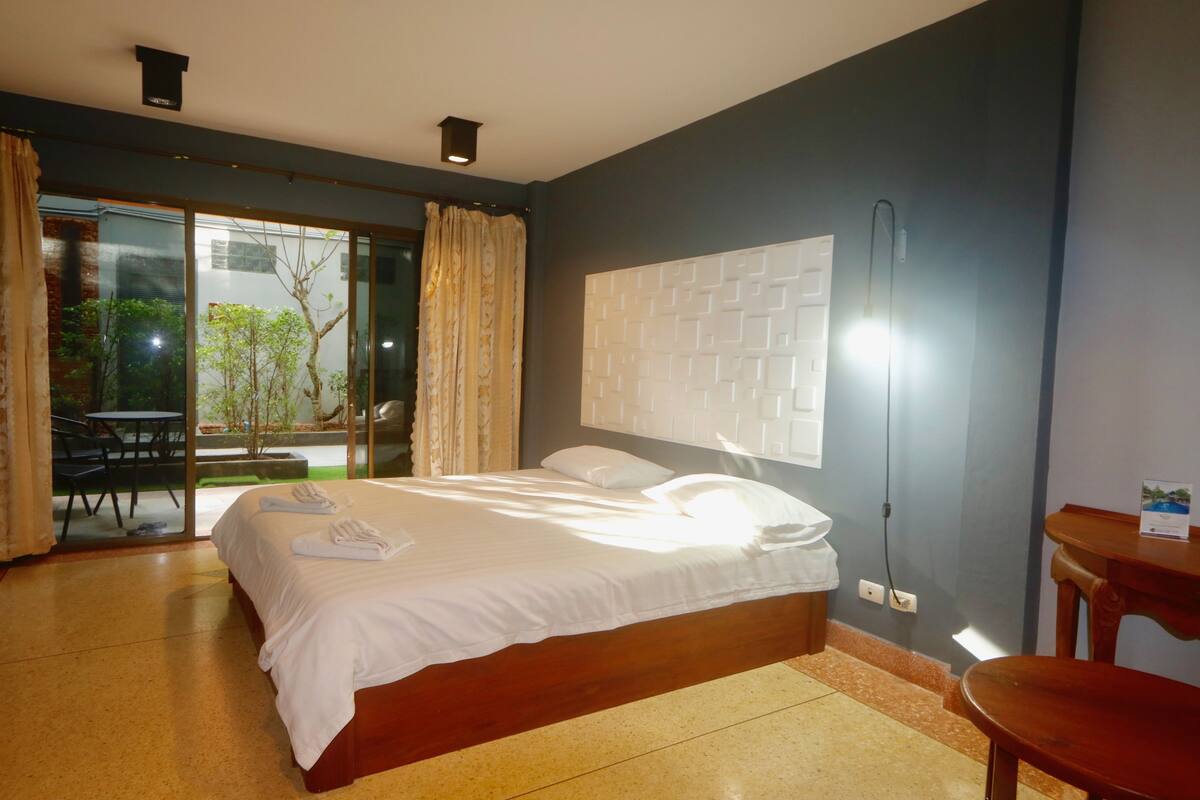 A bedroom with a bed, a nightstand, a window with curtains, and a view of the outdoors.