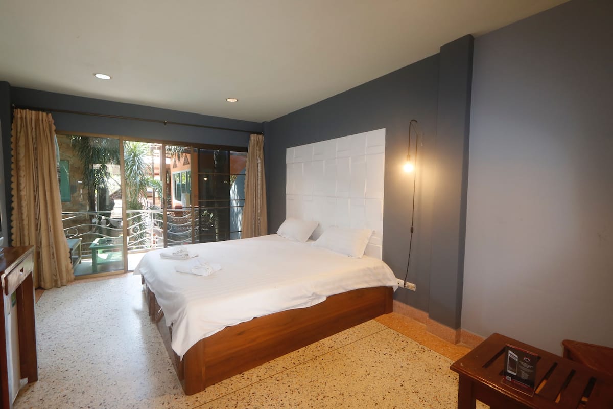 A bedroom with a large bed, a desk, and a chair. There is a balcony with a view of palm trees.