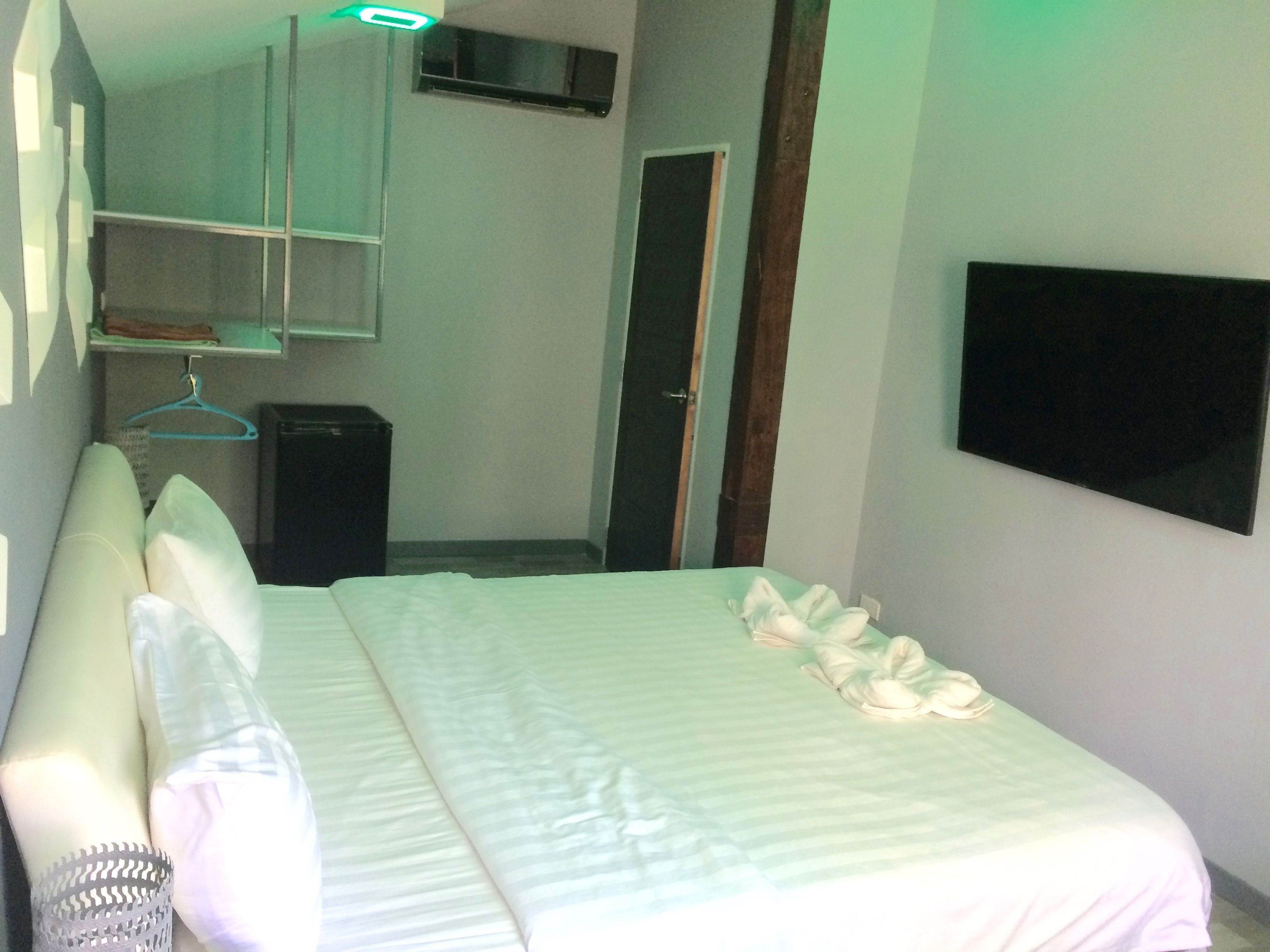 A neatly made bed with pillows and a blanket, a small bedside table, a wall-mounted TV, and a door leading to another room.