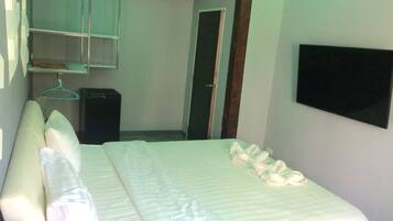 A neatly made bed with pillows and a blanket, a small bedside table, a wall-mounted TV, and a door leading to another room.