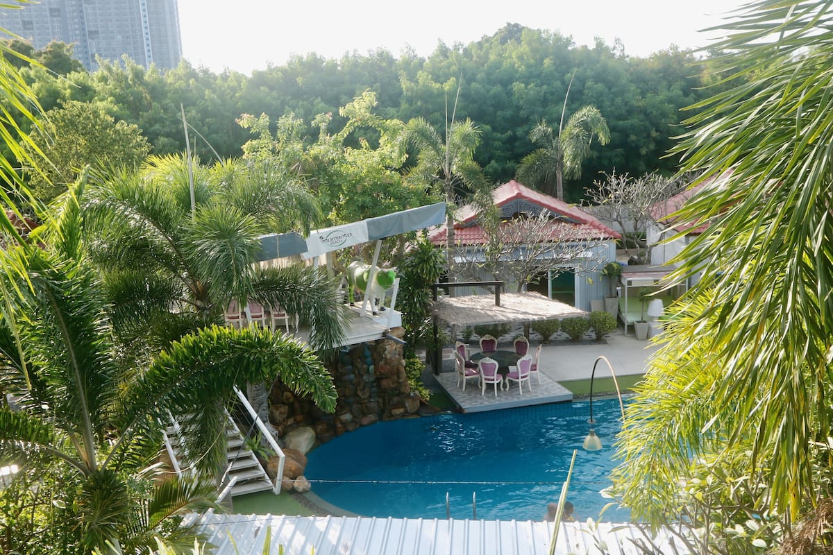 A swimming pool surrounded by lush greenery, a patio area with outdoor seating, and a building with a red roof.