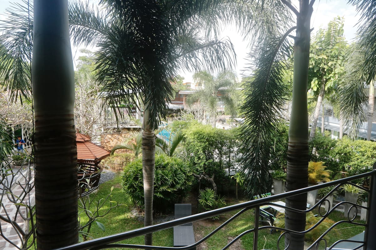 A view from a balcony with a metal railing, overlooking a courtyard with palm trees, a wooden gazebo, and a building in the background.