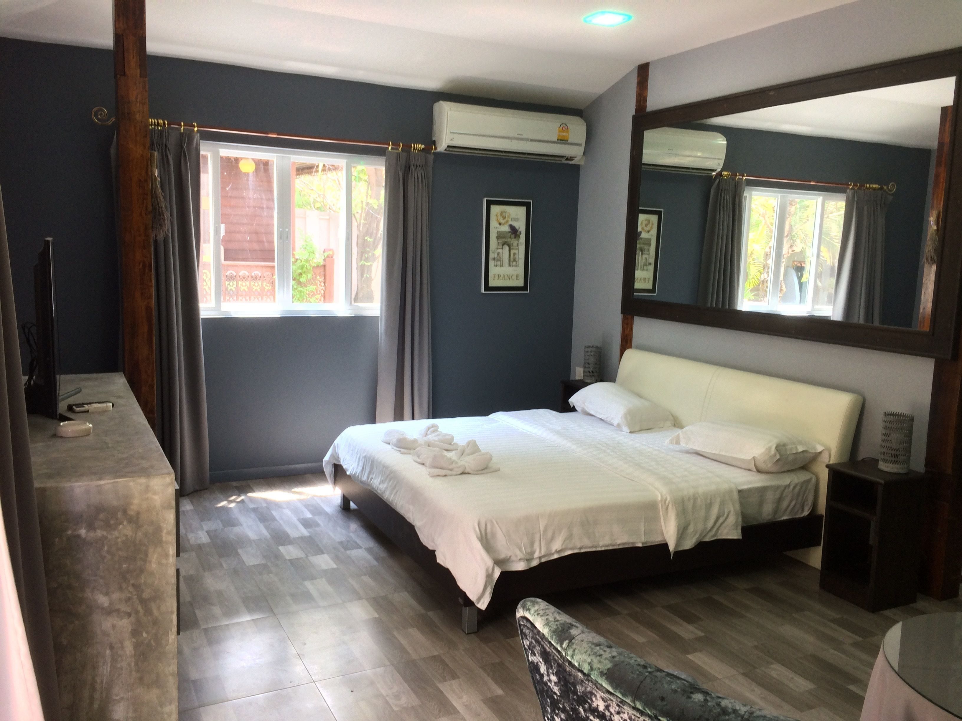A bedroom with a bed, a bedside table, a chair, a large mirror, and an air conditioning unit.