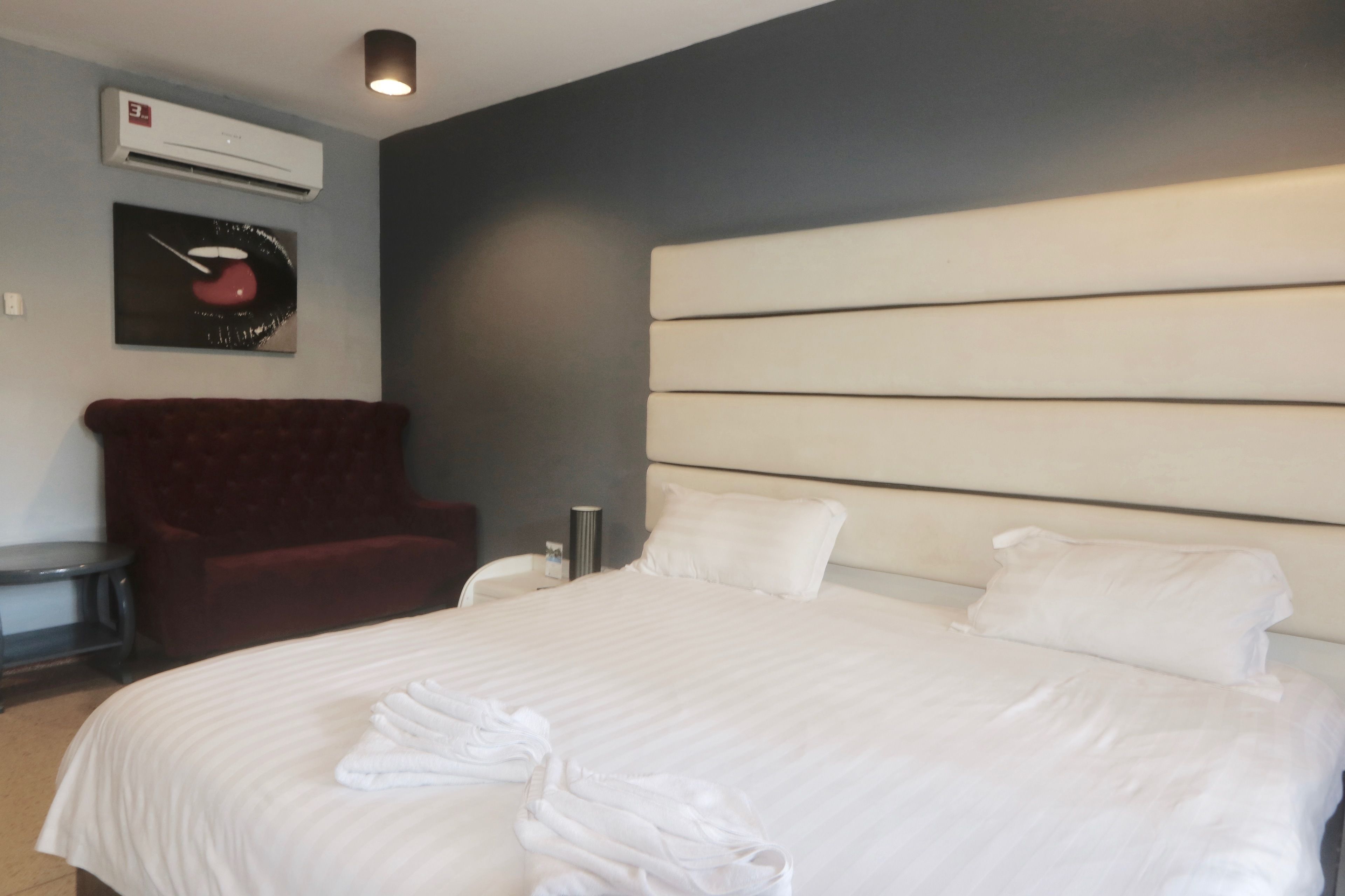 Une chambre d’hôtel avec un grand lit, un fauteuil rouge, un climatiseur fixé au mur et un tableau.