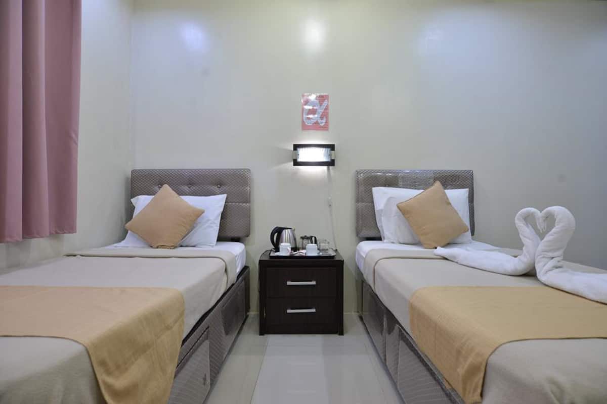 A hotel room with two beds, each with a pillow and a blanket. There is a nightstand between the beds with a kettle and a cup.