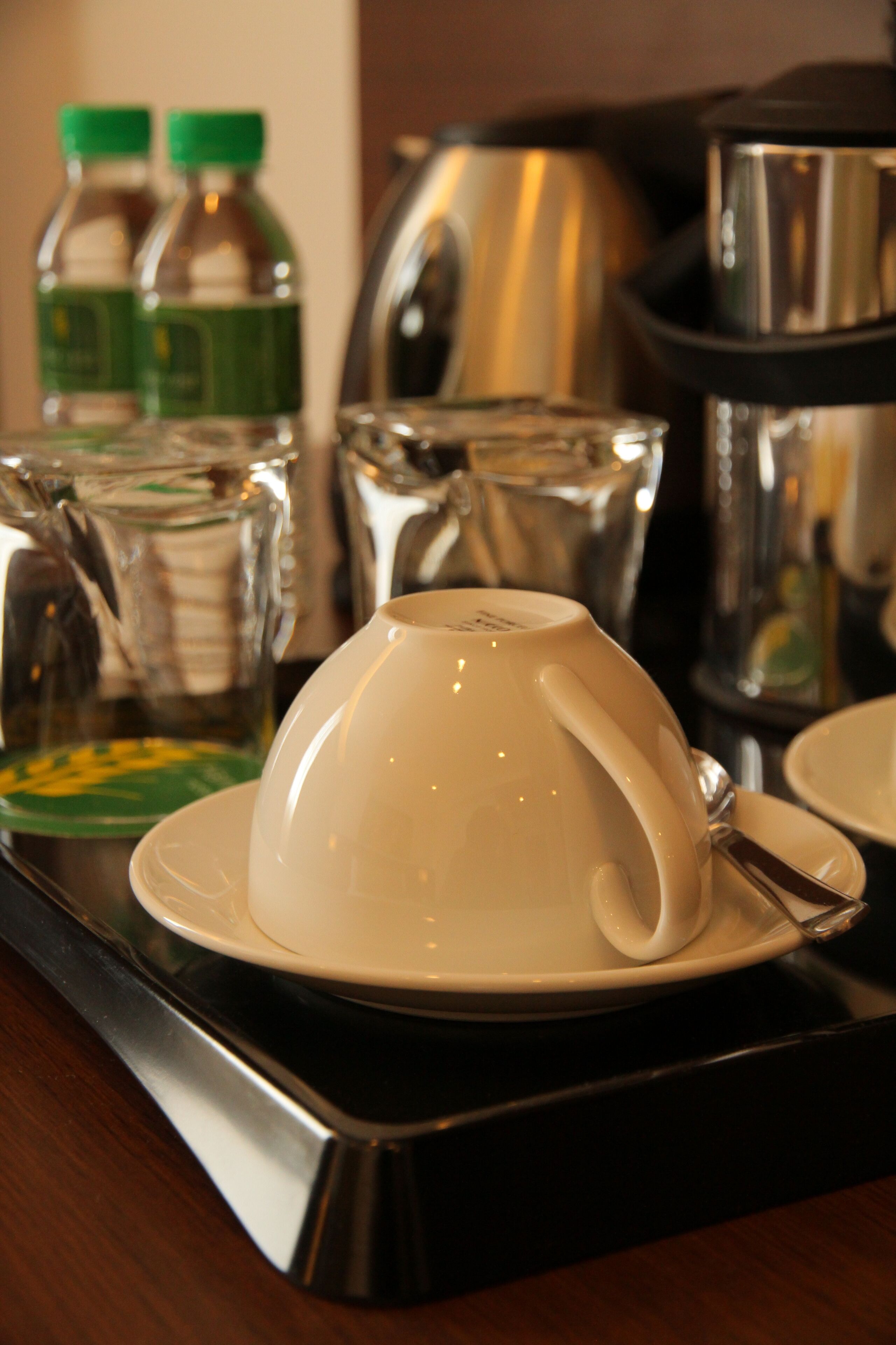 A white cup on a black tray with a spoon, surrounded by kitchen appliances and bottles.