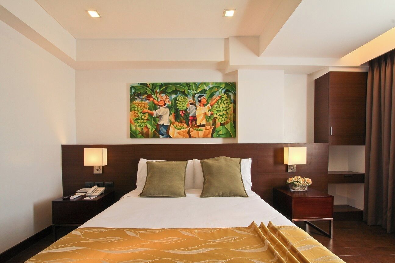 A hotel room with a large bed, two bedside lamps, a nightstand with a phone, and a painting of people holding fruit on the wall.
